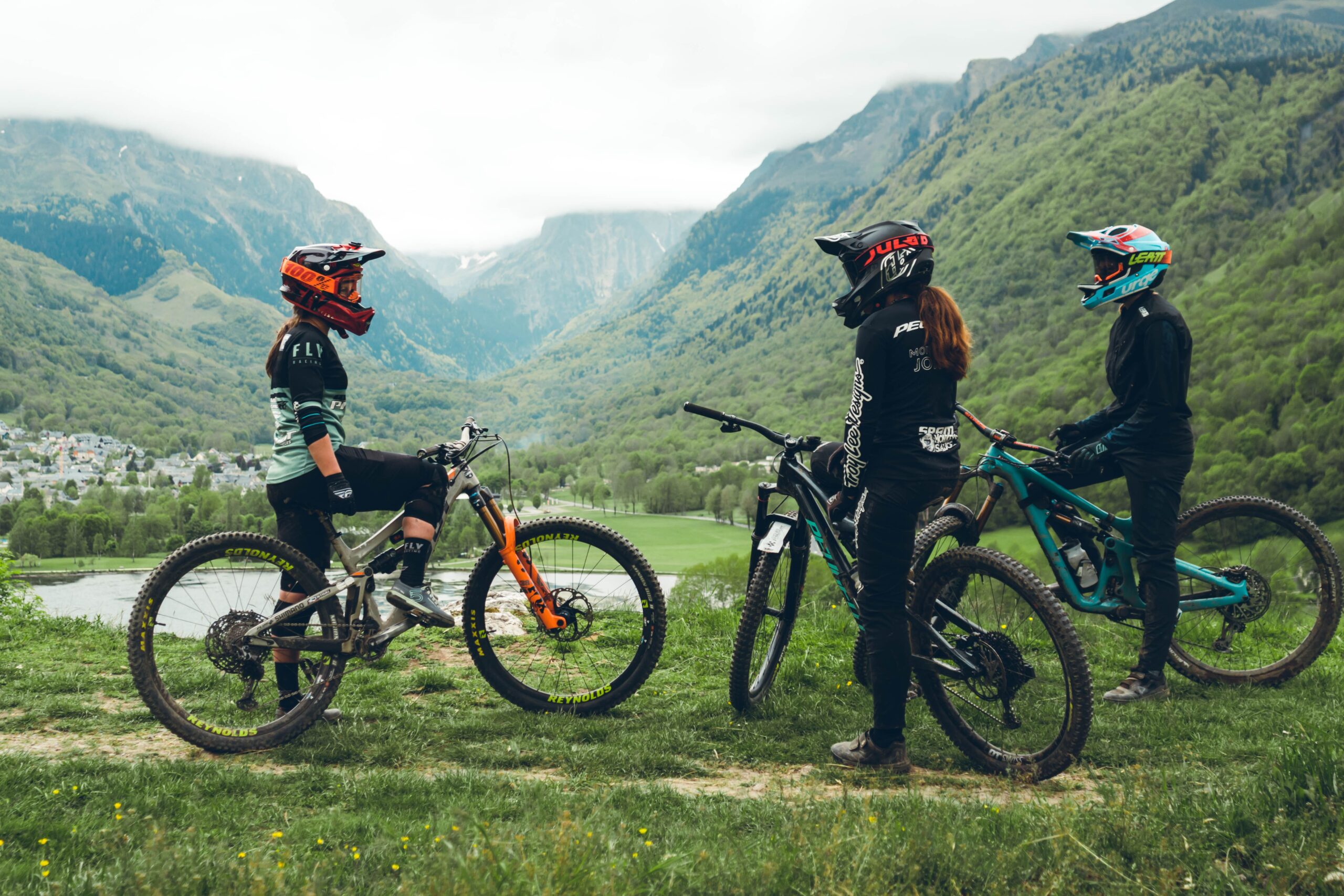 Trois vététistes équipés de protections complètes sur un sentier de montagne dans la vallée de Loudenvielle, Pyrénées, prêts à dévaler une piste de VTT enduro.